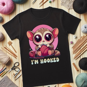 May include: Black t-shirt with a cartoon illustration of a lemur holding knitting needles and yarn. The text "I'M HOOKED" is printed below the lemur.