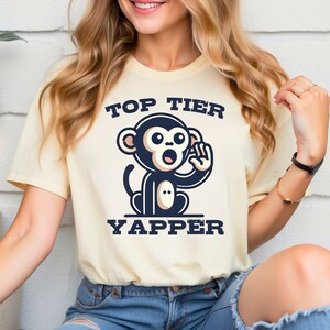 May include: Cream-colored t-shirt featuring a cartoon monkey illustration with the words "TOP TIER YAPPER" in navy blue. The monkey is navy blue and white, with an open mouth and raised hand.