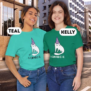 Pu&ograve; includere: T-shirt turchesi e verdi con un gatto bianco con un cappello da mago e una bacchetta. Il testo "MEOWGICAL" &egrave; sotto il gatto. Le modelle indossano jeans blu.