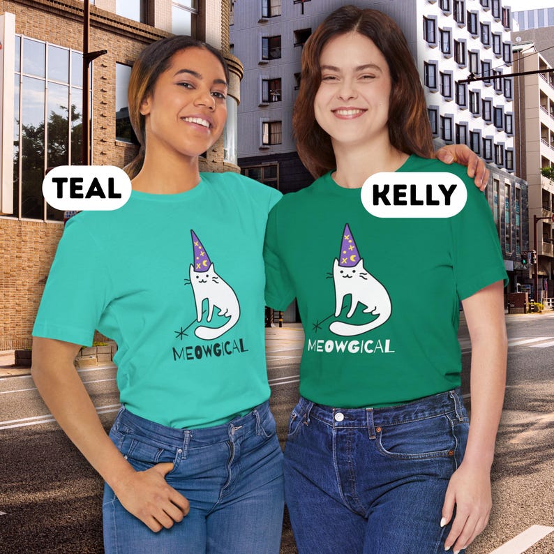 Pu&ograve; includere: T-shirt turchesi e verdi con un gatto bianco con un cappello da mago e una bacchetta. Il testo "MEOWGICAL" &egrave; sotto il gatto. Le modelle indossano jeans blu.