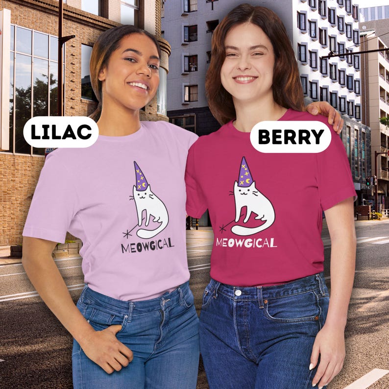 Pu&ograve; includere: Due donne indossano magliette con un disegno di gatto dei cartoni animati. Le magliette sono di colore lilla e bacca, con le parole "LILAC" e "BERRY" in testo bianco sopra le donne. Il gatto &egrave; bianco con un cappello da mago e la parola "MEOWGICAL" &egrave; sotto.