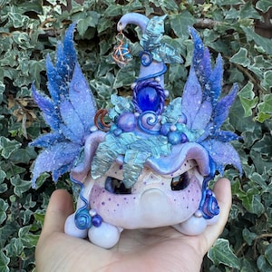 May include: A small, light purple creature figurine with large dark eyes and speckled cheeks. It wears a tall, pointed hat adorned with blue leaves, a central dark blue gemstone, and a dangling star charm. Large, textured blue wings extend from the sides.