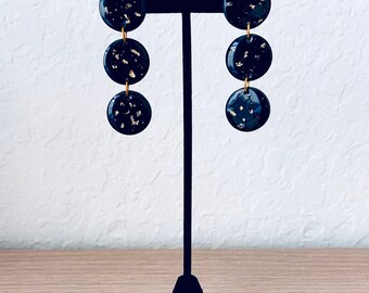 May include: A pair of black and gold dangle earrings with three round discs on each earring. The discs are black with gold flecks.