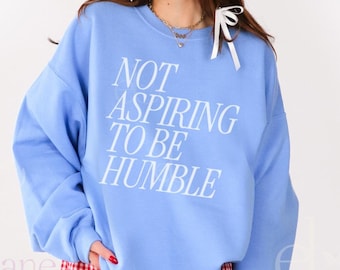 Könnte beinhalten: Ein hellblauer Pullover mit dem Aufdruck "NOT ASPIRING TO BE HUMBLE" in Weiß. Das Model trägt rot-weiß karierte Shorts und schwarze Cowboy-Stiefel.