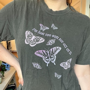 May include: A dark grey t-shirt with a graphic print of butterflies and the text "I'm just too soft for all of it".