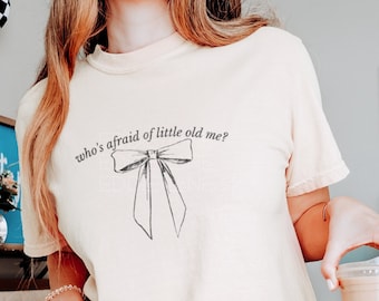 May include: A light yellow cropped t-shirt with a black bow and the text "who's afraid of little old me?" printed on the front. The shirt is being worn by a person with long blonde hair and is paired with light green shorts.