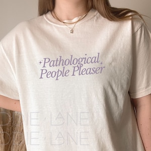 May include: A white oversized t-shirt with the text "Pathological People Pleaser" printed in purple on the front.