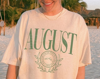 May include: A woman wearing a white baseball cap and sunglasses is standing on a beach. She is wearing a light beige t-shirt with the word "AUGUST" printed in green letters. The t-shirt has a green laurel wreath design with the words "Living for the moments of the good life" printed inside the wreath.