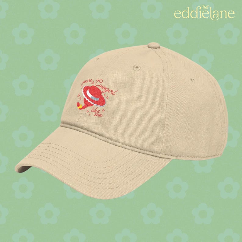 Puede incluir: Una gorra de b&eacute;isbol beige con un gr&aacute;fico bordado de un sombrero de vaquero rojo y el texto "you're a cowgirl like me" en cursiva roja. La gorra tiene una visera curva, un bot&oacute;n en la parte superior y ojales cosidos para la ventilaci&oacute;n.