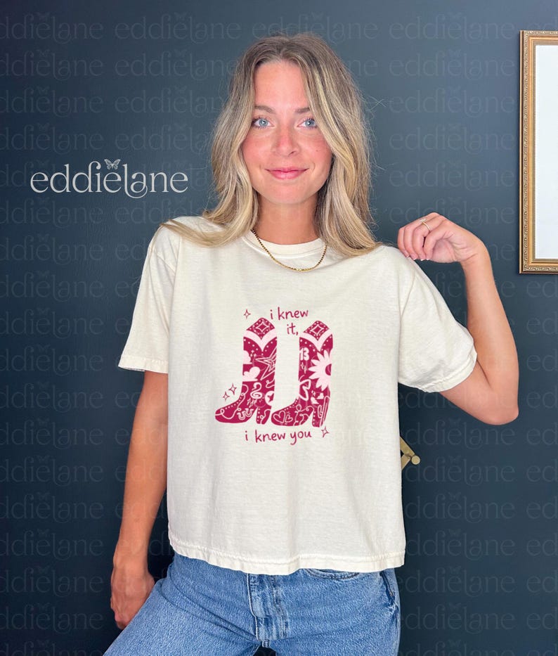 woman wearing an ivory colored comfort colors women's boxy tee with a graphic of 2 cowgirl boots with a bandana design featuring toy story and swiftie inspired doodles