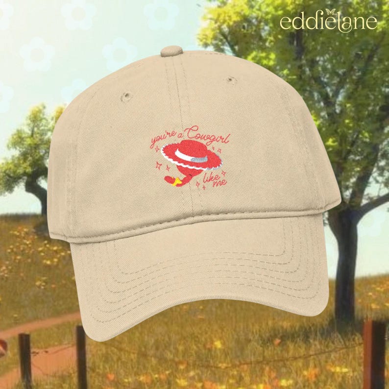 Puede incluir: Una gorra de b&eacute;isbol beige con un gr&aacute;fico bordado de un sombrero de vaquero rojo y el texto "you're a Cowgirl like me" en cursiva roja, acentuado con peque&ntilde;os s&iacute;mbolos de estrellas rojas, centrado en el panel frontal de la corona.