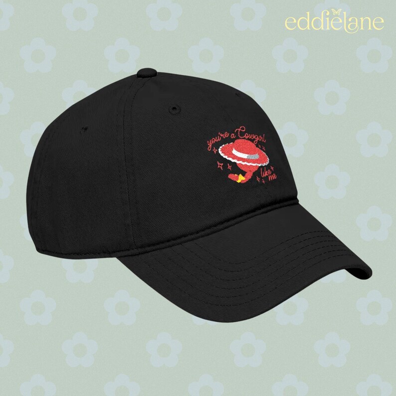 Puede incluir: Una gorra de b&eacute;isbol negra con un gr&aacute;fico bordado de un sombrero de vaquero rojo con una banda blanca y el texto "you're a cowgirl like me" en letra cursiva roja, acentuado por peque&ntilde;as formas de estrellas rojas en el panel frontal.
