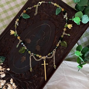 K&ouml;nnte beinhalten: Eine goldfarbene Halskette mit einem Schwertanh&auml;nger, verziert mit gr&uuml;nen Blatt- und Blumenanh&auml;ngern sowie verschiedenen Perlen. Die Halskette wird auf einer dunklen Holzoberfl&auml;che mit einem gr&uuml;n-wei&szlig; karierten Hintergrund pr&auml;sentiert.