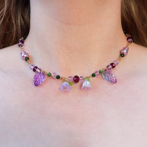 May include: A delicate gold chain necklace with purple and green glass beads and flower charms. The necklace features small purple leaves and bell-shaped flowers.