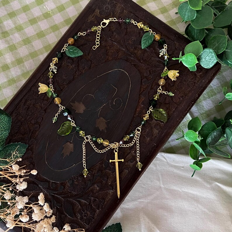 K&ouml;nnte beinhalten: Eine goldfarbene Halskette mit einem Schwertanh&auml;nger, verziert mit gr&uuml;nen Blatt- und Blumenanh&auml;ngern sowie verschiedenen Perlen. Die Halskette wird auf einer dunklen Holzoberfl&auml;che mit einem gr&uuml;n-wei&szlig; karierten Hintergrund pr&auml;sentiert.