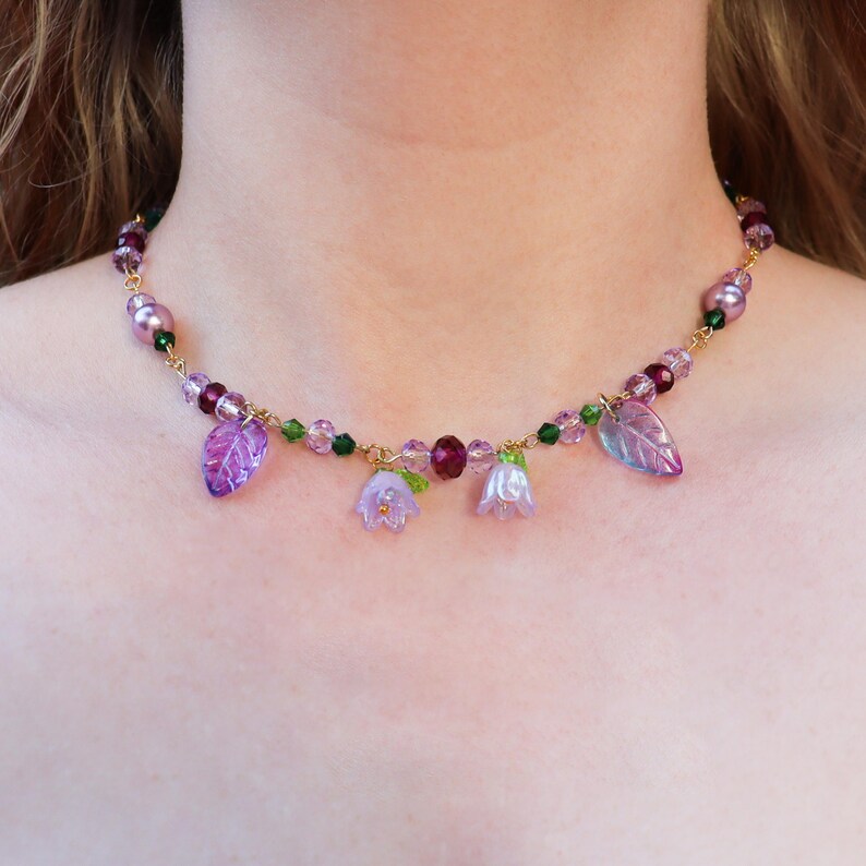 May include: A delicate gold chain necklace with purple and green glass beads and flower charms. The necklace features small purple leaves and bell-shaped flowers.