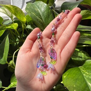 May include: A delicate necklace with a floral theme. It features purple and green glass beads, iridescent leaves, and small flower charms.