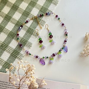 May include: A delicate purple and green necklace and earrings set with flower and leaf charms. The necklace is made with small glass beads and a gold chain.