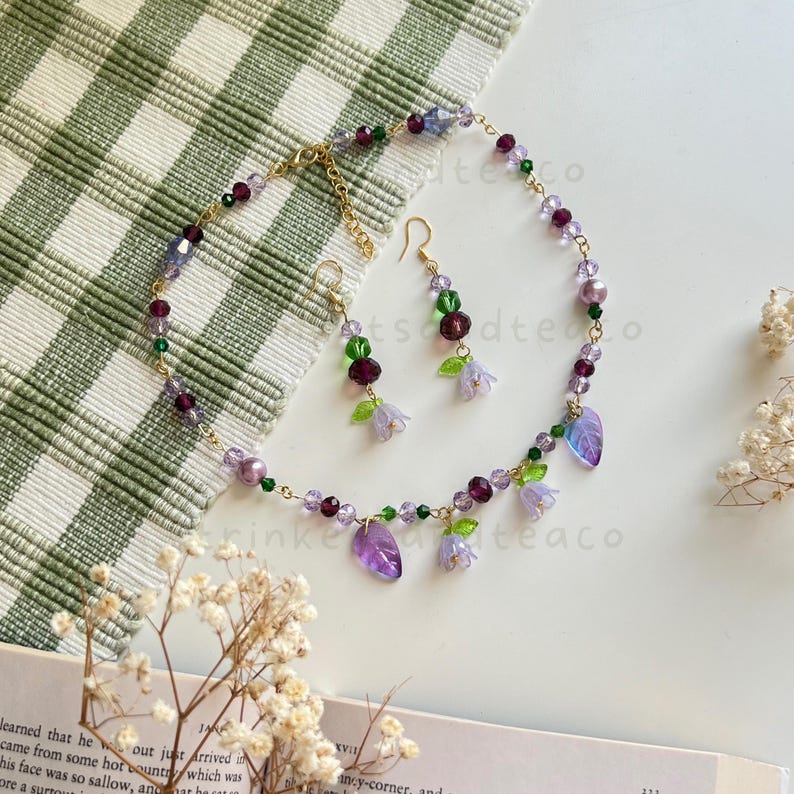 May include: A delicate purple and green necklace and earrings set with flower and leaf charms. The necklace is made with small glass beads and a gold chain.