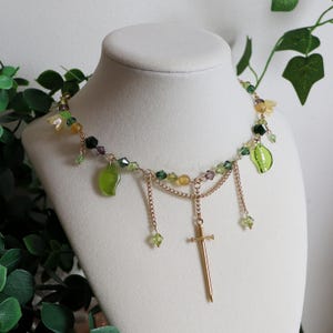 K&ouml;nnte beinhalten: Halskette mit goldfarbener Kette, Schwertanh&auml;nger und gr&uuml;nen Blattanh&auml;ngern. Die Halskette hat gr&uuml;ne, gelbe und violette Perlen mit herabh&auml;ngenden Ketten. Sie wird auf einer wei&szlig;en Schaufensterpuppe pr&auml;sentiert.
