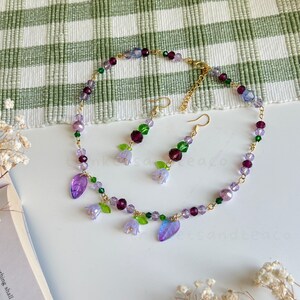 May include: A delicate gold necklace and matching earrings feature purple and green glass beads, small flower charms, and leaf accents.