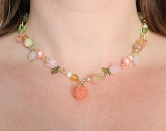 May include: A delicate necklace featuring a gold chain adorned with a variety of beads in pastel colors. The necklace includes pink, green, and yellow beads, along with small flower and leaf charms, and a coral-colored raspberry pendant.