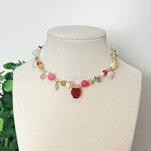 May include: A beaded necklace with a strawberry pendant. The necklace features pink, green, and white beads, with a gold-colored wire. The pendant is a red, textured strawberry. The necklace is displayed on a white mannequin.