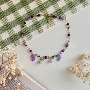 May include: A delicate gold chain necklace with purple, green, and clear glass beads. The necklace features a pendant with three purple flower charms and two green leaf charms.