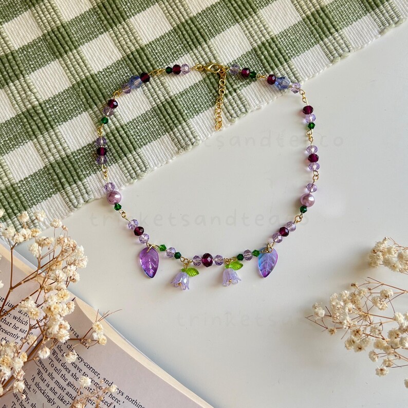 May include: A delicate gold chain necklace with purple, green, and clear glass beads. The necklace features a pendant with three purple flower charms and two green leaf charms.