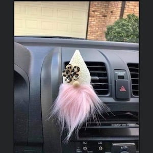 May include: A white and pink gnome car air freshener with a leopard print flower on the hat. The gnome is hanging from the air vent in a car.