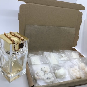 Puede incluir: Una caja de cartón marrón con una tapa dorada está abierta, revelando una botella de vidrio transparente con una tapa dorada y una cadena dorada. La caja también contiene varias pequeñas fundentes de cera blancas en bolsas de plástico transparentes.