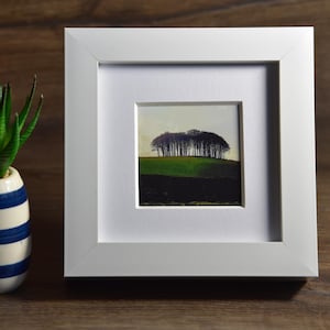 May include: A framed landscape art print depicting trees on a green hill under a pale sky. A small potted succulent with green leaves is to the left. The pot is white with blue stripes.