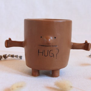 Pode incluir: Caneca de cerâmica marrom com braços e pernas, um rosto e o texto "HUG?" na frente.