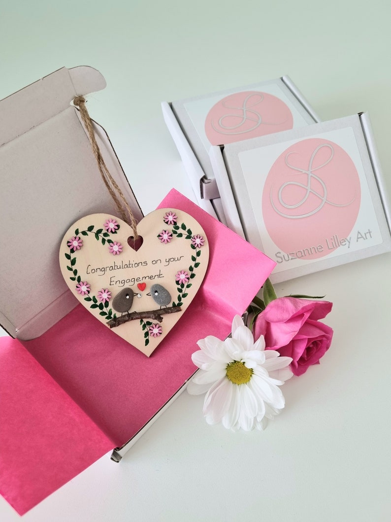 Handmade engagement pebble art heart with two pebble birds perched on a real twig branch, pink and white hand-painted flowers, gem accent, and "Congratulations on your engagement" wording.