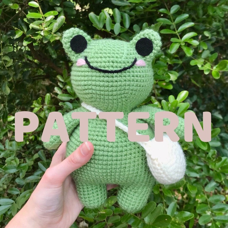 May include: Green crocheted frog plush toy with black eyes, a white bag, and a pink smile. The frog is holding the bag in its right hand. The text "PATTERN" is written in pink letters across the image.