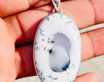 May include: Oval pendant with a white stone featuring black speckles and a gray center, set in a silver-colored metal frame. The pendant hangs from a silver chain, creating a striking contrast.