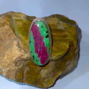 May include: Oval ring with a ruby-zoisite stone. The stone displays a central band of deep pink-red, framed by bright green. The ring's setting is silver-toned, with a detailed, textured border. The ring is a unique piece of jewellery.