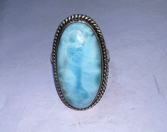 Op de afbeelding: Ovale ring met een lichtblauwe edelsteen, omlijst door een zilverkleurige, gedraaide touwachtige rand. De edelsteen heeft een wolkachtig uiterlijk met tinten blauw en wit. De ring is gezet op een zilveren band, een klassiek sieraadontwerp.