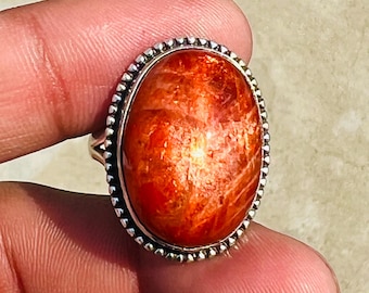 May include: A silver ring with an oval-shaped sunstone. The sunstone is a deep orange color with a shimmery, iridescent effect. The ring has a beaded edge.