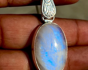 Op de afbeelding: Zilveren hanger met een ovale, iriserende maansteen. De steen heeft een blauwe en witte gloed. De hanger is bevestigd aan een zilveren ketting. De bovenkant van de hanger heeft een sierlijk ontwerp.