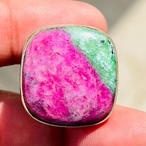 Könnte beinhalten: Ein quadratischer Ring mit einem polierten, mehrfarbigen Edelstein, der einen leuchtend magentafarbenen Bereich und einen kontrastierenden hellgrünen Bereich aufweist, eingefasst in eine schlichte, glatte silberfarbene Metallfassung.