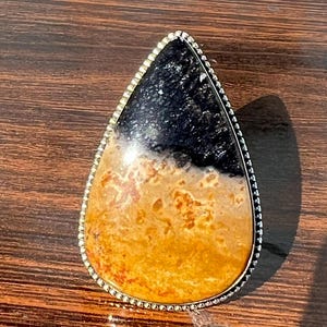 Könnte beinhalten: Ein tropfenförmiger Anhänger mit einem schwarzen und orangefarbenen Achat-Stein, der in einer silbernen Fassung mit einem perlenbesetzten Rand gefasst ist.