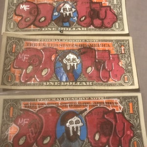 Puede incluir: Tres billetes de un dólar estadounidense con las palabras "MF DOOM" escritas en graffiti rojo, naranja y negro en cada billete. Los billetes están ligeramente desgastados y tienen un aspecto vintage.