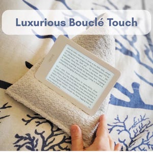 May include: A Kindle e-reader with a cream-colored screen is displayed on a folded, light beige bouclé towel. The text "Luxurious Bouclé Touch" is at the top. The e-reader is held by a hand.