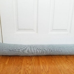 Puede incluir: Un tapón de puerta gris hecho de tela, colocado en la parte inferior de una puerta blanca.