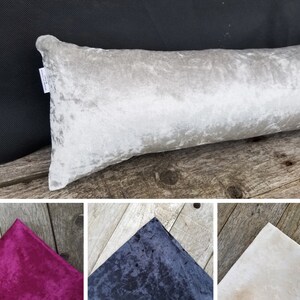 Può includere: Un cuscino lungo in velluto bianco con un leggero luccichio. Il cuscino è appoggiato su una superficie di legno.