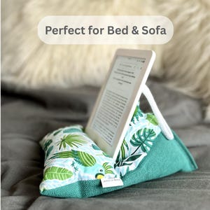 May include: A teal and white reading pillow with a tropical leaf pattern, supporting a white e-reader. The pillow has a triangular shape with a white rope support and a small tag that says "Made in Greece".