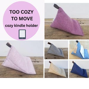 May include: A collection of cozy Kindle holders in various colors, including purple, gray, and blue. Each holder is triangular, made of soft fabric, and features a small handle. The image includes text that reads "TOO COZY TO MOVE cozy kindle holder."
