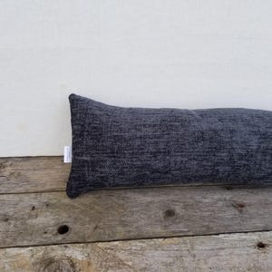 Può includere: Un paraspifferi rettangolare grigio scuro in tessuto strutturato. Il lungo cuscino cilindrico poggia su una superficie di legno stagionato, appoggiato a un muro bianco. Una piccola etichetta è attaccata al lato.