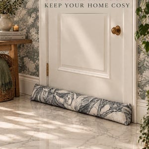 Puede incluir: Un tope de puerta cilíndrico y largo de tela con un patrón monocromático de toile de jouy con querubines y follaje. El texto BLOCK THE DRAUGHT • KEEP YOUR HOME COSY está impreso en la puerta blanca sobre el tope de tela para la puerta.